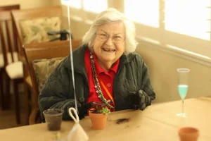 An elderly woman wearing glasses and a jacket sits at a table, smiling, with a small potted plant and soil in front of her.