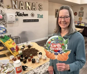 A smiling woman stands in a kitchen with gardening supplies on the counter, holding a sign that reads, "The rotation of the Earth really makes my day.