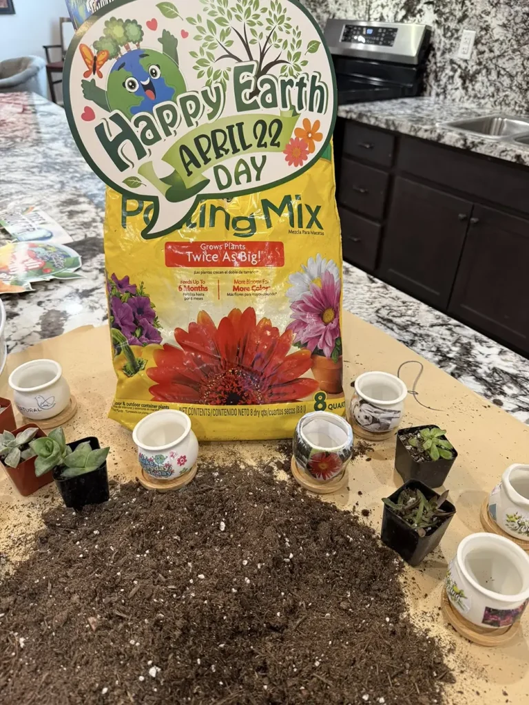 A bag of potting mix labeled "Happy Earth Day April 22" stands behind several small potted succulents and overturned soil on a kitchen countertop.
