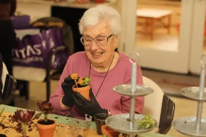 An elderly woman wearing glasses and gloves smiles while arranging a small potted succulent at a table with other plants and gardening materials.
