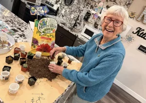 An elderly woman smiles while planting small pots with soil on a kitchen counter. A bag labeled "Happy Earth Day Potting Mix" and various small plants are visible.