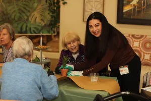 A woman assists an elderly woman with planting a small potted plant at a table, while two other elderly women sit nearby.