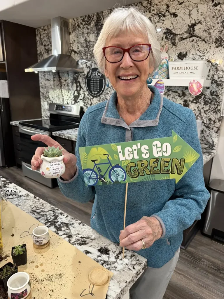 An older woman with gray hair and glasses smiles while holding a small potted succulent and a sign that says &ldquo;Let&rsquo;s Go Green&rdquo; in a kitchen with a granite countertop.