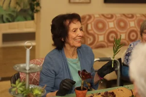 An elderly woman wearing gloves smiles while holding a plant cutting and sitting at a table with potted plants.