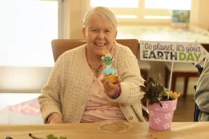 An elderly woman smiles while holding a decorated cupcake. A potted plant and a sign reading "Go planet, it's your Earth Day" are in the background.