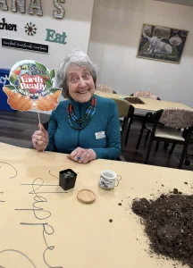 Older woman seated at a table holding a sign that says "The rotation of the farm really makes my day," with a small potted plant, teacup, and spilled soil on the table.