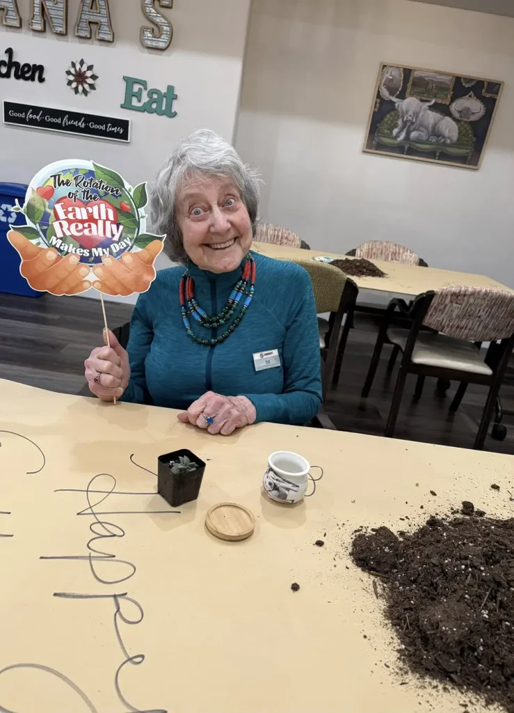 Older woman seated at a table holding a sign that says "The rotation of the farm really makes my day," with a small potted plant, teacup, and spilled soil on the table.