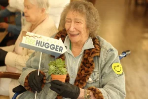 An elderly woman smiles while holding a potted succulent and a sign that reads "tree HUGGER." She is seated and wearing a denim jacket with a smiley face patch on the sleeve.
