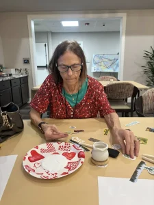 A woman wearing glasses and a red patterned top sits at a table arranging stickers near a white ceramic plate with heart designs and painting supplies.