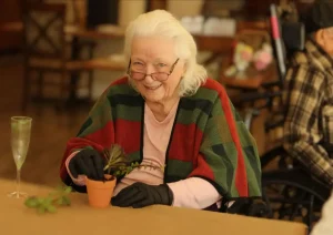 An elderly woman wearing glasses and gloves smiles while potting a small plant at a table, with another person in a wheelchair in the background.