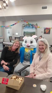 Two women sit at a table next to a person in an Easter Bunny costume, surrounded by festive decorations and snacks, enjoying fun Easter activities in a bright room.