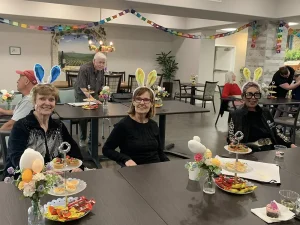 A group of older adults wearing bunny ears sit at decorated tables with food and holiday-themed centerpieces, enjoying Easter activities in a festive indoor setting.