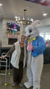 An elderly woman stands next to a person in an Easter Bunny costume in a living room. She uses a walker, and there is a TV and holiday decorations in the background, suggesting she's enjoying some Easter activities with loved ones.