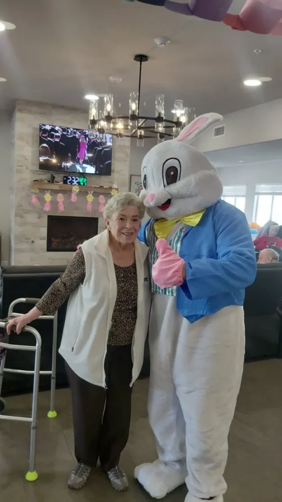 An elderly woman stands next to a person in an Easter Bunny costume in a living room. She uses a walker, and there is a TV and holiday decorations in the background, suggesting she's enjoying some Easter activities with loved ones.