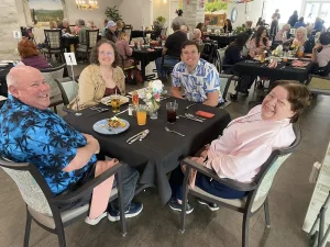 Four people sit at a table in a busy restaurant, smiling at the camera as they enjoy Easter activities together. Plates of food and drinks are on the table, with other diners visible in the background.