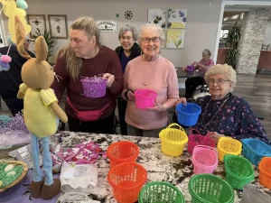 Four elderly women sit and stand around a table with colorful plastic baskets, shredded paper, and a bunny decoration, getting ready to enjoy Easter activities together.