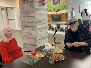 An elderly woman in a red dress and a man in a wheelchair sit at a table with snacks and candy, enjoying Easter crafts in a warm communal indoor setting.