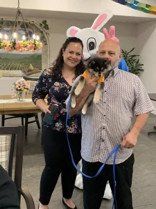 A man and woman pose with a small dog on a leash as the Easter Bunny stands behind them in a decorated indoor setting, capturing the joy of festive Easter activities.