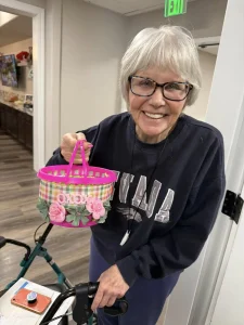 An older woman with glasses and gray hair smiles while holding a small pink and plaid basket decorated with pink flowers—perfect for Easter activities. She is wearing a dark sweatshirt and standing indoors.