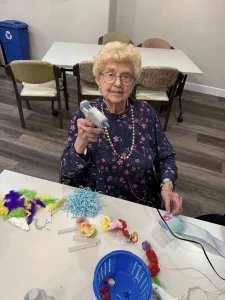 An elderly woman sitting at a table holds a hot glue gun, surrounded by colorful craft supplies such as feathers, flowers, and yarn—perfect for creative Easter activities.