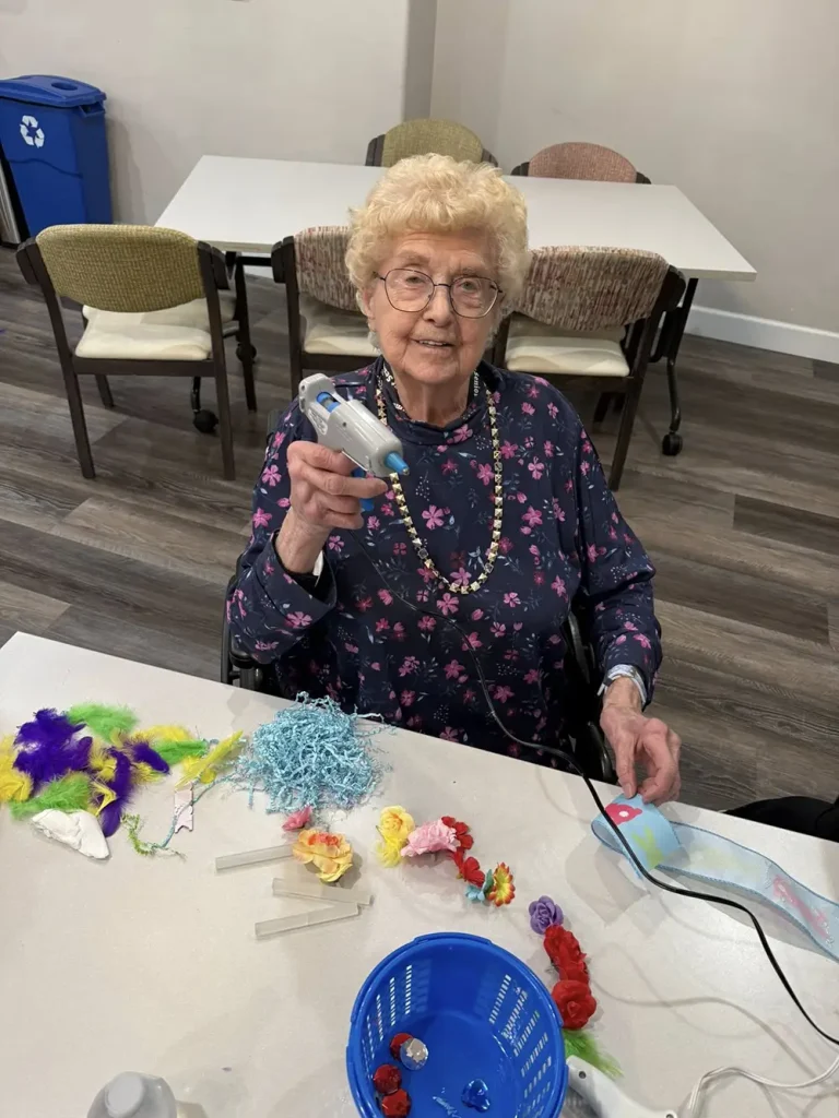 An elderly woman sitting at a table holds a hot glue gun, surrounded by colorful craft supplies such as feathers, flowers, and yarn&mdash;perfect for creative Easter activities.