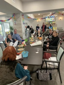 A group of people sit around a decorated table at a festive indoor gathering, enjoying Easter activities. Some wear bunny ear headbands, with party decorations hanging in the background.
