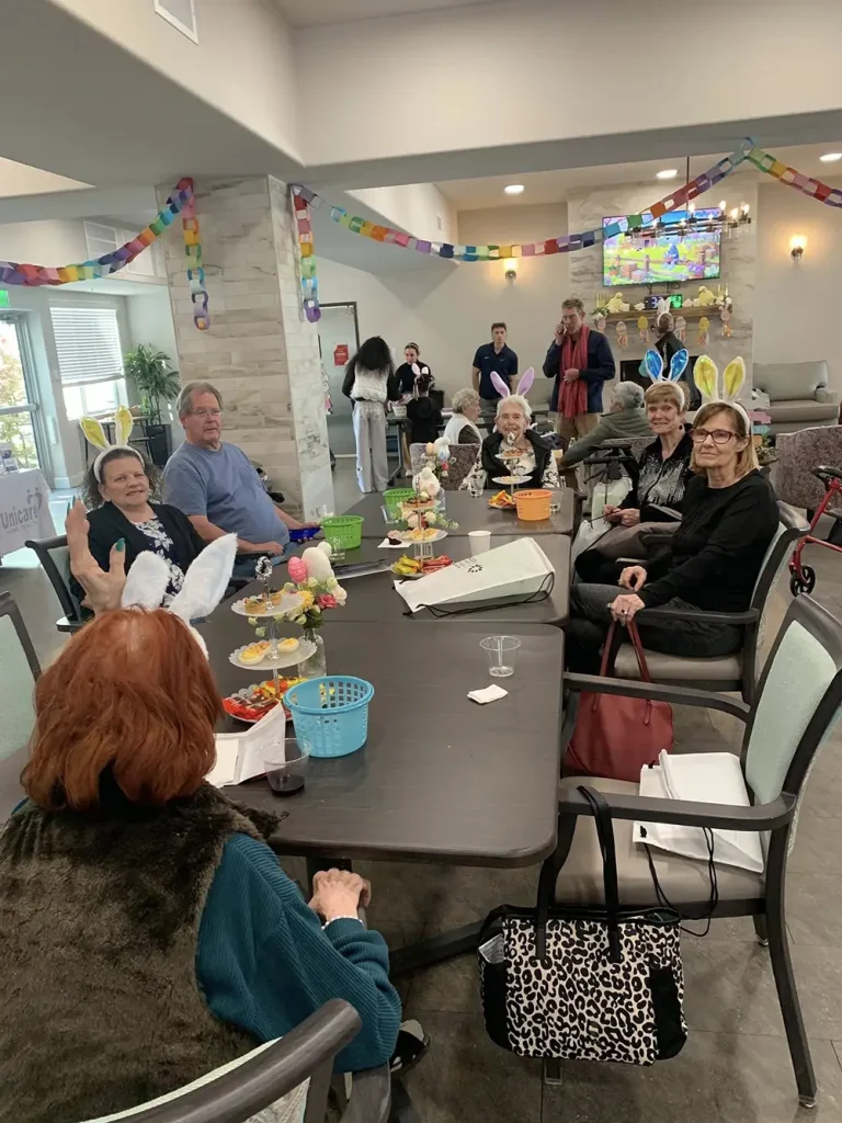 A group of people sit around a decorated table at a festive indoor gathering, enjoying Easter activities. Some wear bunny ear headbands, with party decorations hanging in the background.