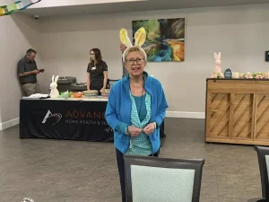 An older woman wearing bunny ears stands in a room decorated for Easter, with a food table, people in the background, and Easter games adding to the festive atmosphere.