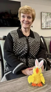 An older woman with short light hair sits and smiles at a table, enjoying Easter activities, with a small yellow Easter bunny decoration in front of her.