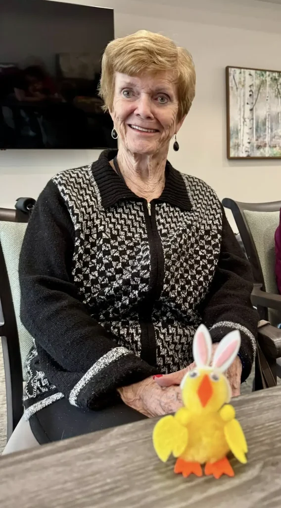An older woman with short light hair sits and smiles at a table, enjoying Easter activities, with a small yellow Easter bunny decoration in front of her.