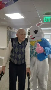 An elderly man with a walker poses next to a person in an Easter Bunny costume giving a thumbs up in a hallway, sharing smiles during Easter activities.