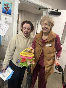 An elderly woman with a walker stands next to a younger woman holding a colorful basket decorated with flowers. Both are indoors and appear to be interacting as they enjoy Easter activities together.