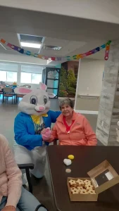 A person in an Easter Bunny costume sits beside an older woman at a table with cookies and eggs, enjoying Easter activities; colorful paper chains decorate the room.