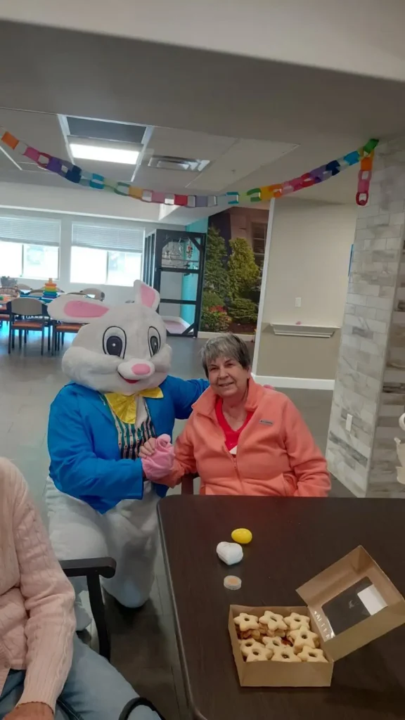 A person in an Easter Bunny costume sits beside an older woman at a table with cookies and eggs, enjoying Easter activities; colorful paper chains decorate the room.