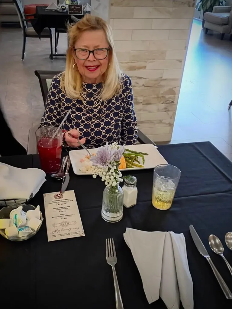 A woman with glasses sits at a table set for elevated dining, holding a fork as she enjoys her meal. The elegant black tablecloth, flowers, drinks, utensils, and menu complete the sophisticated atmosphere.