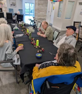 Four elderly people sit around a decorated table with black tablecloth, candles, and flowers, enjoying snacks in a bright communal dining area that showcases elevated dining at its finest.