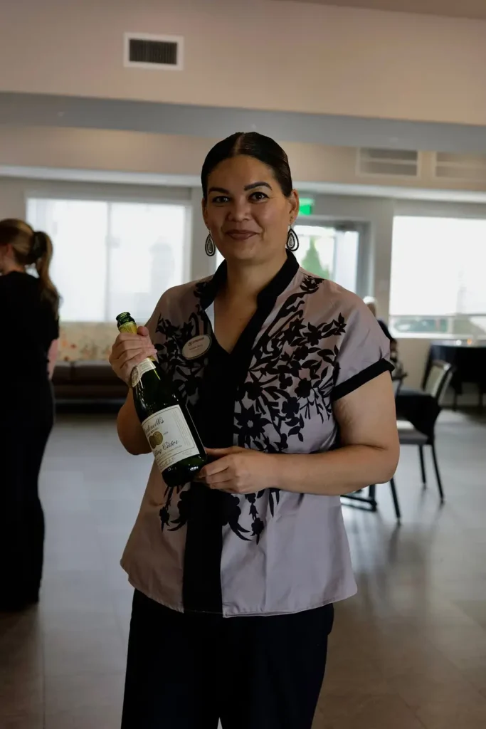 Woman standing indoors holding a bottle of sparkling cider, wearing a name tag and patterned blouse, with tables and chairs in the background&mdash;ready to enjoy an Elevated Dining experience.