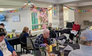 Elderly residents are seated at decorated tables in a dining area, enjoying meals in an Elevated Dining experience. Colorful paper garlands hang from the ceiling and a floral mural brightens the wall.