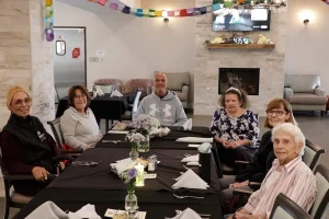 Six older adults sit around a dining table in a decorated room, enjoying elevated dining with folded napkins, flowers, and neatly arranged tableware as they face the camera.