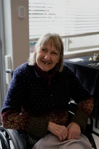 An older woman with gray hair sits in a wheelchair indoors, smiling at the camera. She is wearing a patterned sweater, enjoying the warmth of elevated dining in a welcoming atmosphere.