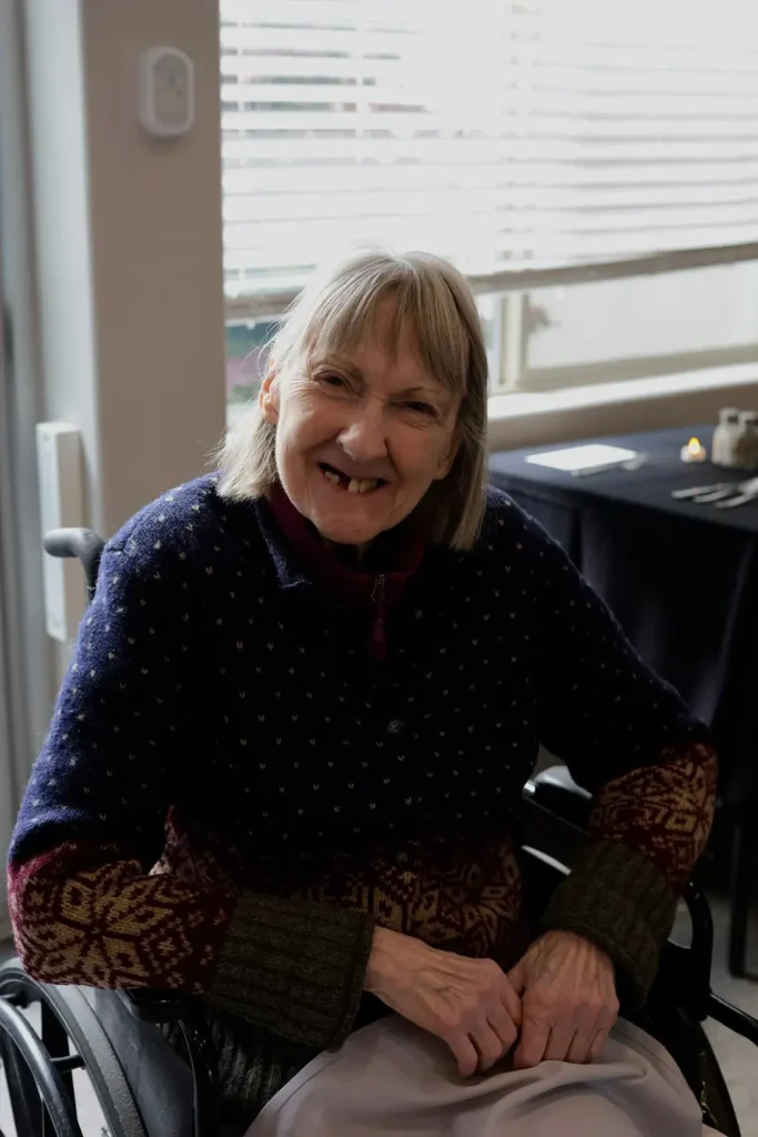 An older woman with gray hair sits in a wheelchair indoors, smiling at the camera. She is wearing a patterned sweater, enjoying the warmth of elevated dining in a welcoming atmosphere.
