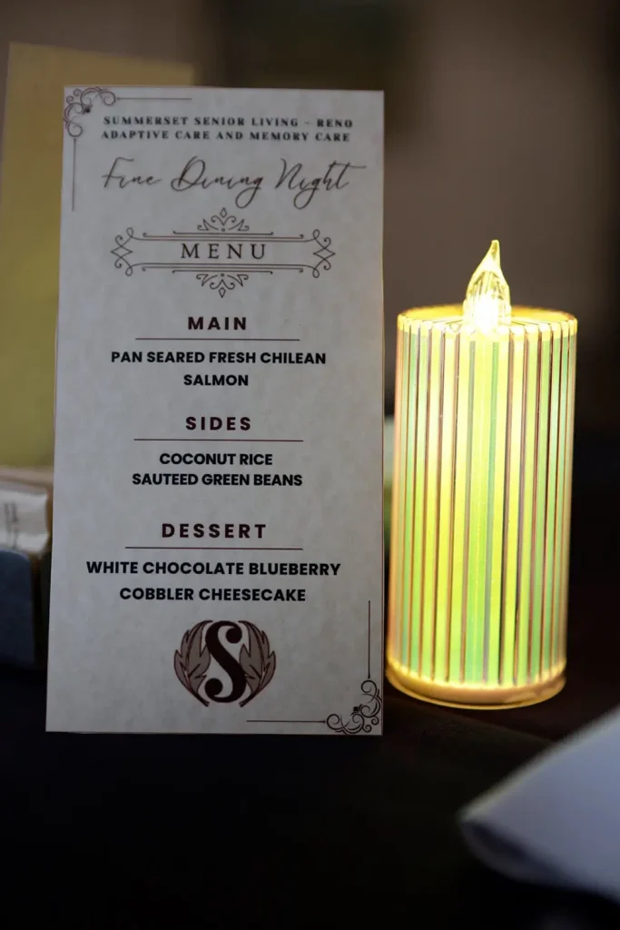 A printed dinner menu featuring salmon, coconut rice, saut&eacute;ed green beans, and cheesecake promises an elevated dining experience beside a lit electric candle on a dark surface.