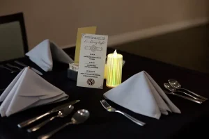 A formal table setting with folded white napkins, cutlery, a menu card, and a lit candle on a dark tablecloth creates an elevated dining experience.