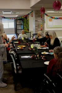 A group of elderly women sit around a long table with food and drinks, decorated with flowers and garlands, enjoying an elevated dining experience in a festive indoor setting.