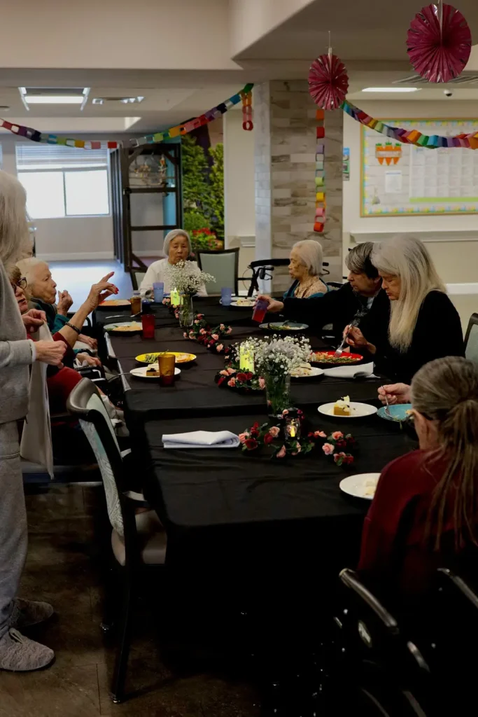 A group of elderly women sit around a long table with food and drinks, decorated with flowers and garlands, enjoying an elevated dining experience in a festive indoor setting.
