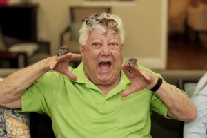 An older man in a green shirt sits on a couch with several small frogs on his head and hands, smiling with his mouth open as if enjoying an Exotic Animal Safari right at home.
