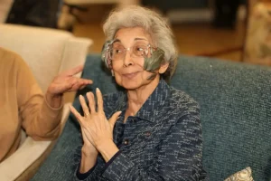 Elderly woman with gray hair and glasses sits on a blue couch, wearing curved plastic barriers attached to each side of her glasses, resembling protective gear for an animal safari.