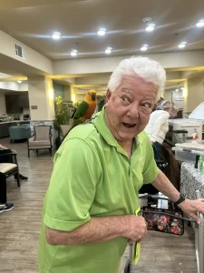 An elderly man in a green polo shirt uses a walker and has a colorful parrot perched on his shoulder, bringing an Exotic Animal Safari vibe to the well-lit communal room.