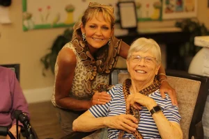 Two women smiling indoors on an Exotic Animal Safari; one stands with her arm around the seated other, both with a snake draped around their shoulders.