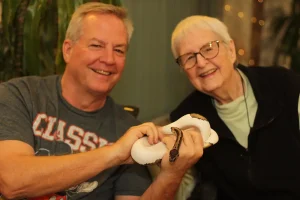 Two smiling people sit indoors during an Exotic Animal Safari, one holding a white and brown snake with both hands while the other person looks on.